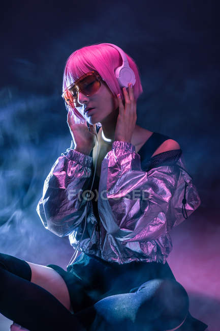Молодая стильная азиатская женщина с фиолетовыми волосами слушает музыку в наушниках, сидя на полу на темном фоне дымки — стоковое фото