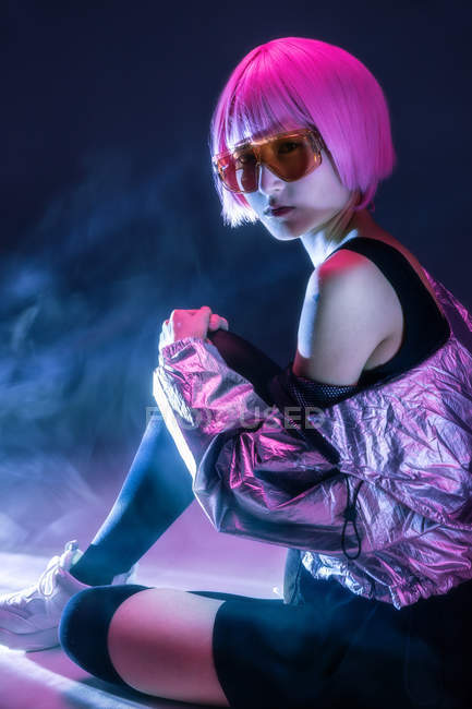 Trendy young Japanese woman with purple hair in silver jacket and red sunglasses sitting on dark haze background — Stock Photo