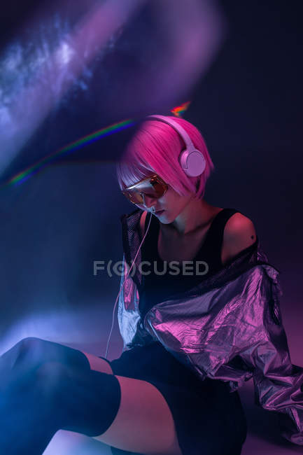 Joven mujer asiática elegante con el pelo púrpura escuchar música con auriculares mientras está sentado en el suelo sobre fondo de neblina oscura - foto de stock