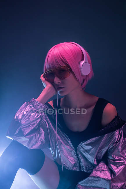 Young stylish Asian woman with purple hair listening music with headphones while sitting on floor on dark haze background — Stock Photo