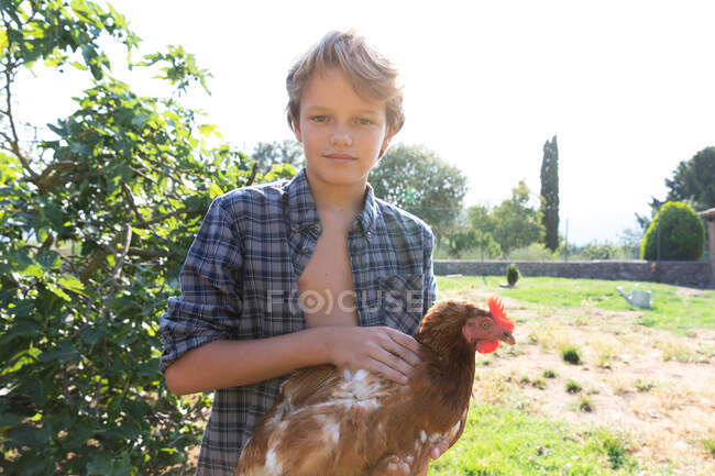 Teen boy and in checkered shirt and denim short petting hen while standing near green bushes on sunny day on farm — Stock Photo