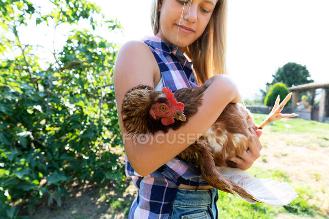 Menina adolescente e em camisa quadriculada e ganga galinha de estimação curta enquanto está perto de arbustos verdes no dia ensolarado na fazenda — Fotografia de Stock