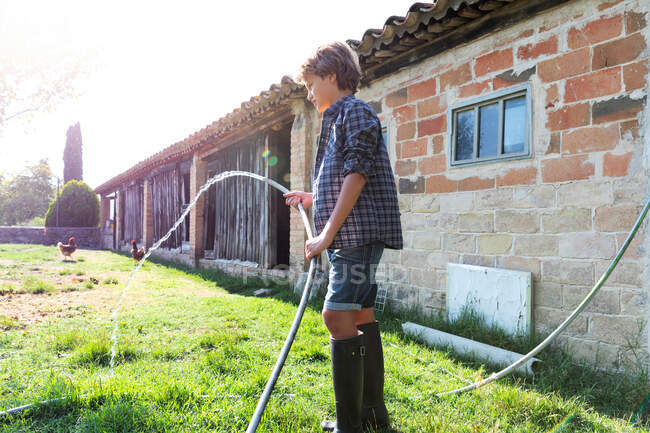 Side view of male teenager in checkered shirt using hose to watering green lawn near brick barn on sunny day on farm — Stock Photo