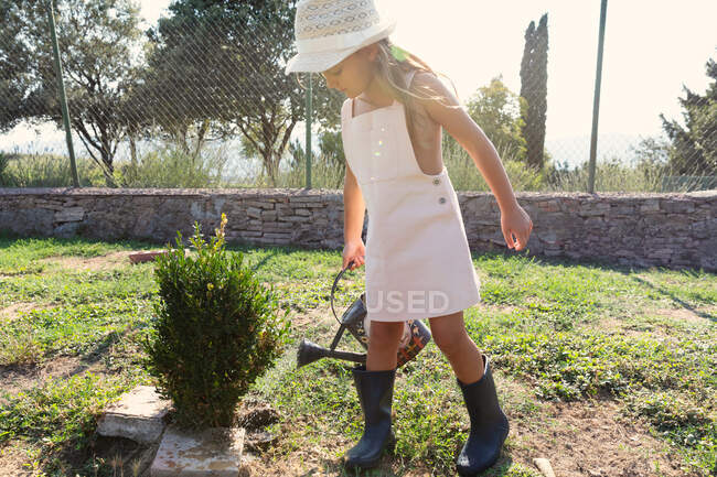 Menina em vestido e chapéu regar pequeno arbusto enquanto ajuda no jardim no dia ensolarado na fazenda — Fotografia de Stock