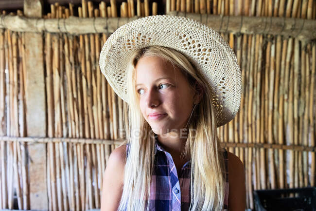 Mujer adolescente en sombrero de paja sonriendo y mirando hacia otro lado mientras está de pie contra la partición de madera en el interior cobertizo en la granja - foto de stock