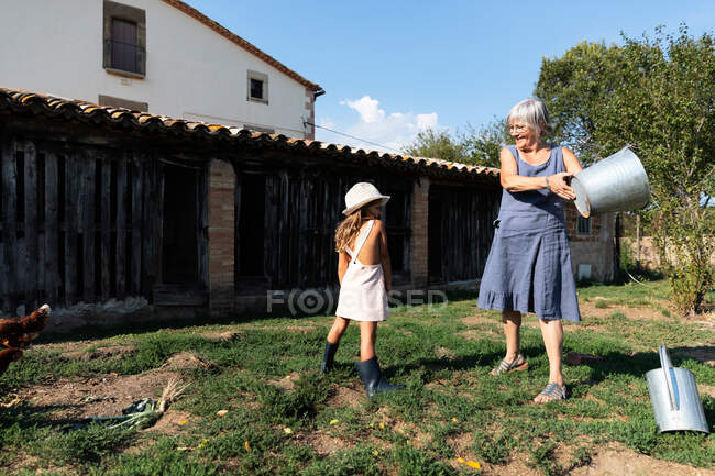 Großmutter spritzt Wasser aus Eimer auf Rasen, während sie an sonnigem Tag neben Enkeln auf der Ranch steht — Stockfoto