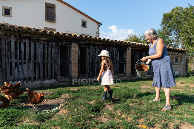Grandmother splashing water from bucket on lawn while standing near grandchildren on sunny day on ranch — Stock Photo