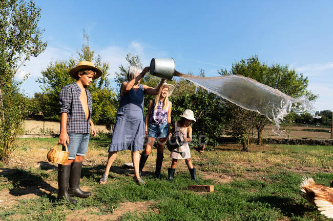 Großmutter spritzt Wasser aus Eimer auf Rasen, während sie an sonnigem Tag neben Enkeln auf der Ranch steht — Stockfoto