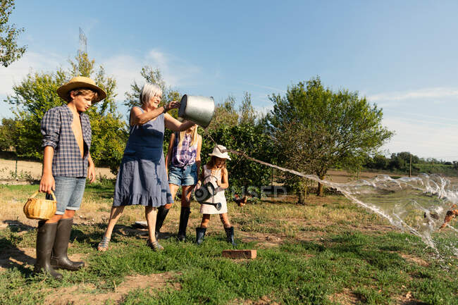 Avó salpicando água do balde no gramado enquanto estava perto de netos no dia ensolarado no rancho — Fotografia de Stock