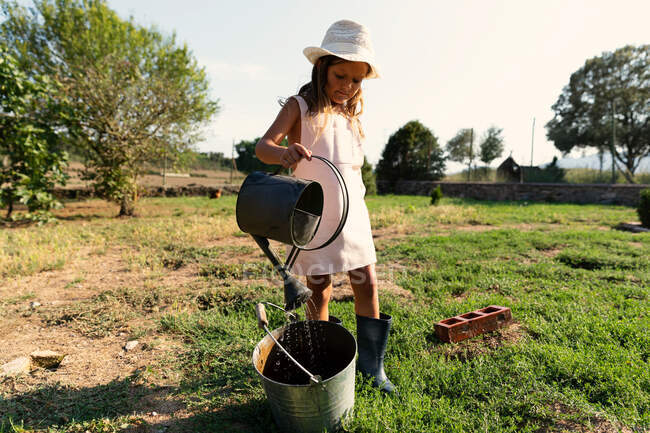 Niña en vestido y sombrero vertiendo agua limpia de lata en cubo mientras está de pie en el patio de la granja en el día soleado - foto de stock