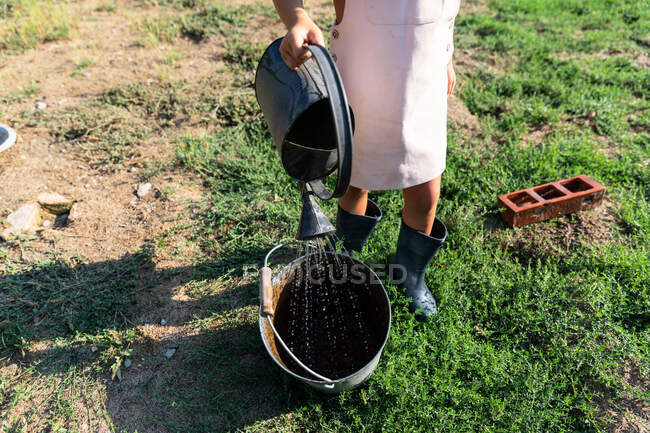 Маленькая девочка в платье и шляпе льет чистую воду из банки в ведро, стоя на ферме в солнечный день — стоковое фото