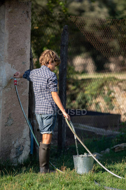 Menino derramando água limpa em balde enquanto estava em pé no quintal no dia ensolarado — Fotografia de Stock