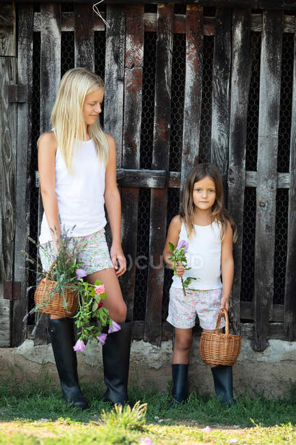 Dos chicas en trajes casuales sonriendo y de pie cerca de granero de madera gruñón mientras pasa tiempo en la granja - foto de stock