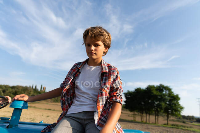 Хлопчик в повсякденному вбранні дивиться на камеру, сидячи на синьому тракторі проти хмарного неба в сонячний день на фермі — стокове фото