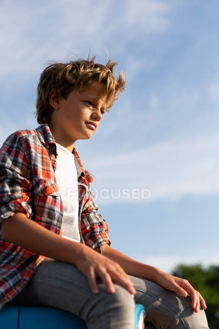 Vista lateral del niño en traje casual mirando hacia otro lado mientras está sentado en el tractor azul contra el cielo nublado en el día soleado en la granja - foto de stock