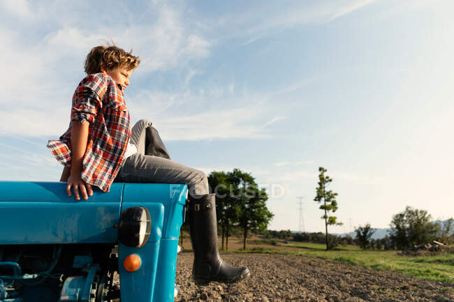 Vista lateral do menino em roupa casual olhando para longe enquanto sentado no trator azul contra o céu nublado no dia ensolarado na fazenda — Fotografia de Stock
