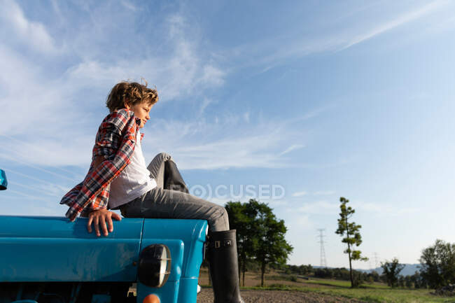 Side view of boy in casual outfit looking away while sitting on blue tractor against cloudy sky on sunny day on farm — Stock Photo