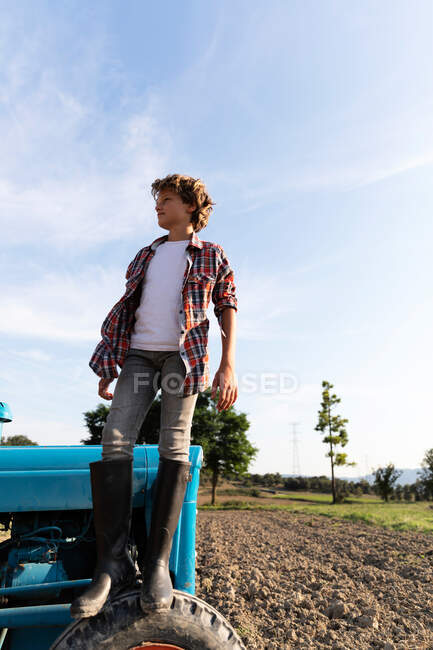 Боковой вид мальчика в повседневной одежде, смотрящего в сторону, стоя на синем тракторе против облачного неба в солнечный день на ферме — стоковое фото