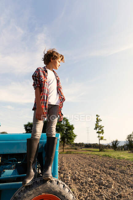 Vista laterale del ragazzo in abito casual guardando lontano mentre in piedi sul trattore blu contro cielo nuvoloso nella giornata di sole in fattoria — Foto stock