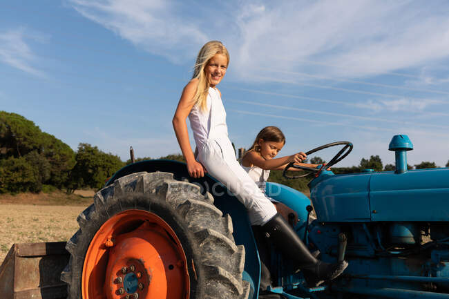 Vue latérale des filles heureuses en tenues décontractées conduisant tracteur bleu sur le terrain agricole par une journée ensoleillée à la ferme — Photo de stock