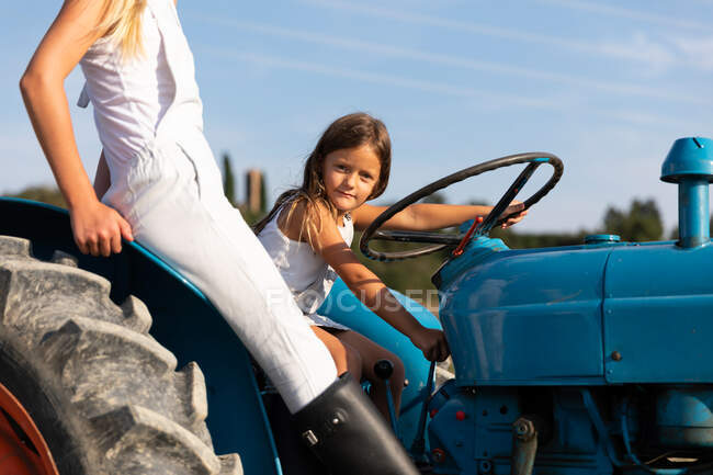 Vue latérale des filles heureuses en tenues décontractées conduisant tracteur bleu sur le terrain agricole par une journée ensoleillée à la ferme — Photo de stock