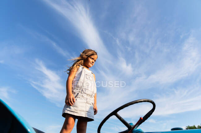 Маленька дівчинка в джинсовій сукні стоїть на тракторі проти хмарного неба на фермі — стокове фото