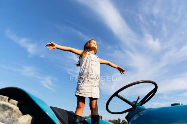 Little girl in denim dress stretching out arms while standing on tractor against cloudy sky on farm — Stock Photo
