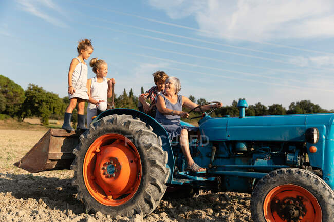 Mujer mayor conduciendo tractor y mirando a los niños mientras trabaja en el campo de agricultura en el día soleado en la granja - foto de stock