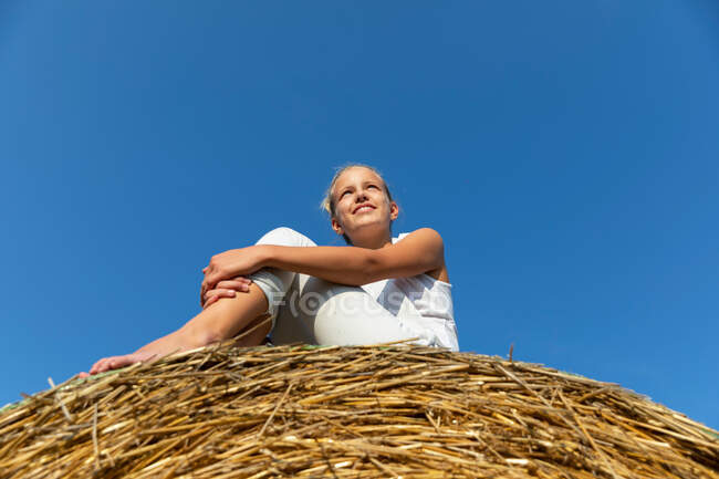 Petite fille regardant loin tout en s'asseyant sur le rouleau d'herbe sèche contre ciel bleu sans nuages sur une journée ensoleillée à la ferme — Photo de stock
