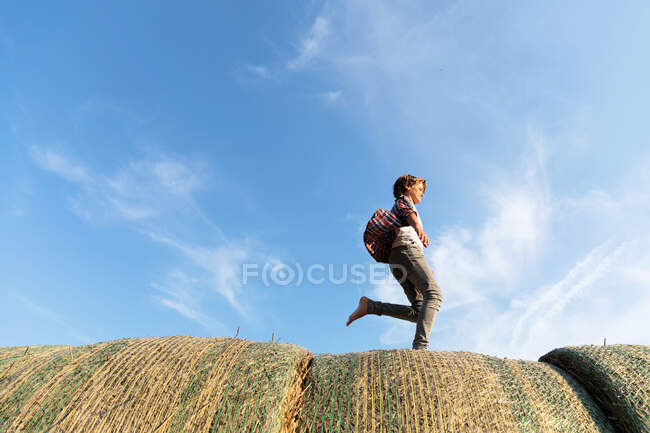 Vista laterale del ragazzo scalzo che corre su rotoli di erba secca contro il cielo blu nuvoloso nella giornata di sole in fattoria — Foto stock