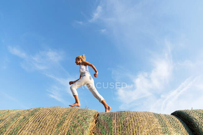 Босоногая девушка, бегущая по рулонам сухой травы на фоне облачного голубого неба в солнечный день на ферме — стоковое фото