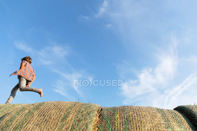 Seitenansicht eines barfüßigen Jungen, der an einem sonnigen Tag auf einem Bauernhof auf Rollen aus getrocknetem Gras gegen bewölkten blauen Himmel läuft — Stockfoto