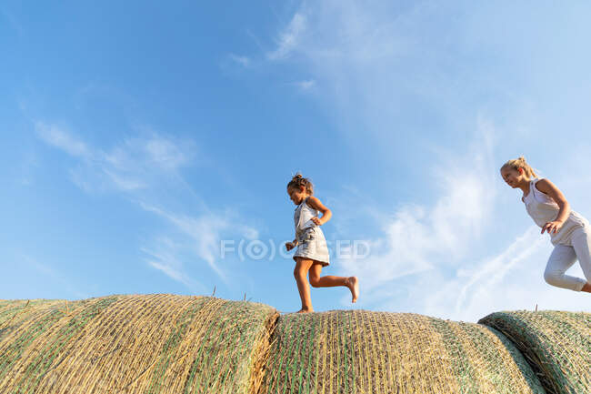 Вид сбоку на трех детей, бегущих вместе по рулонам соломы на фоне облачного голубого неба в поле культуры — стоковое фото