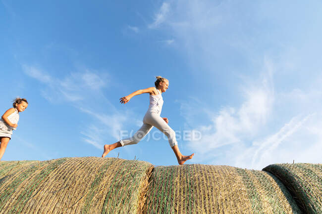 Vista lateral de três crianças correndo em rolos de palha juntos contra o céu azul nublado no campo agrícola — Fotografia de Stock