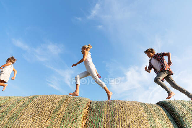 Vista lateral de três crianças correndo em rolos de palha juntos contra o céu azul nublado no campo agrícola — Fotografia de Stock