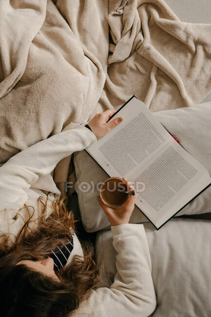 Mulher irreconhecível sentada na cama lendo um livro e bebendo café — Fotografia de Stock