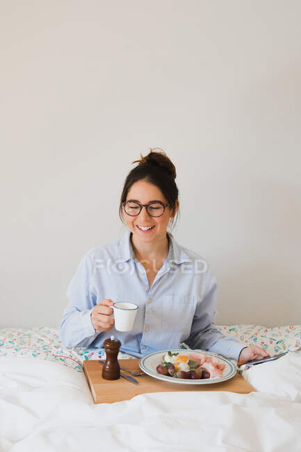 Портрет веселої жінки, що сидить на ліжку з чашкою в руках і підносять здорову їжу на ногах при використанні смартфона — стокове фото