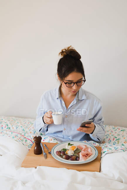 Porträt einer fröhlichen Frau, die mit einer Tasse in der Hand und einem Tablett mit gesundem Essen auf den Beinen im Bett sitzt, während sie ein Smartphone benutzt — Stockfoto