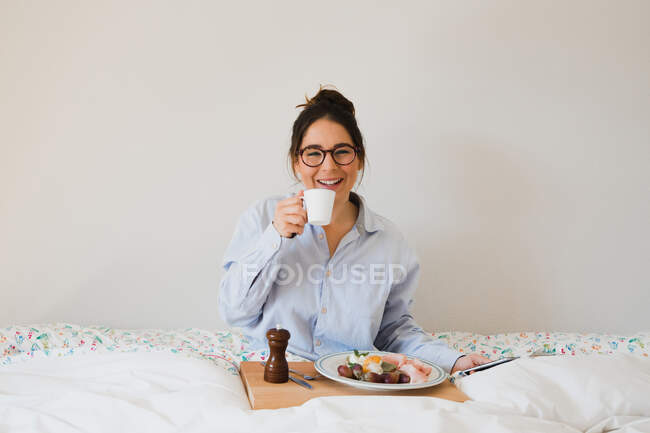 Ritratto di donna allegra seduta sul letto con tazza in mano e vassoio con cibo sano sulle gambe durante l'utilizzo di uno smartphone — Foto stock