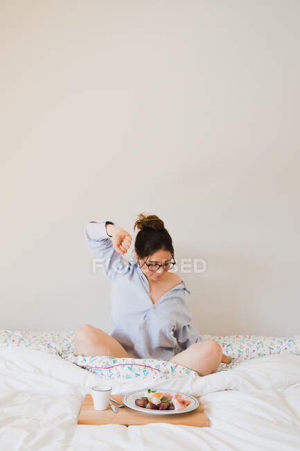 Retrato de mujer estirándose mientras está sentada en la cama frente a una bandeja con comida saludable - foto de stock