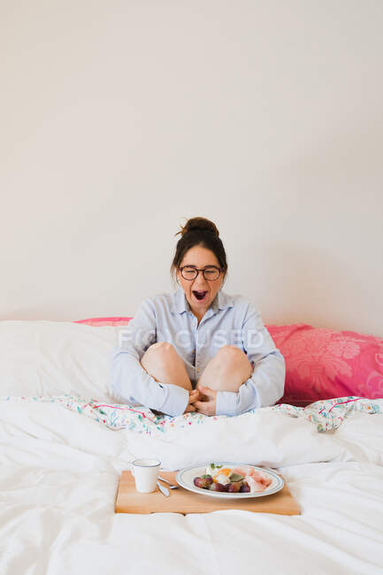 Portrait of woman yawning while sitting on bed in front of a tray with healthy food — Stock Photo