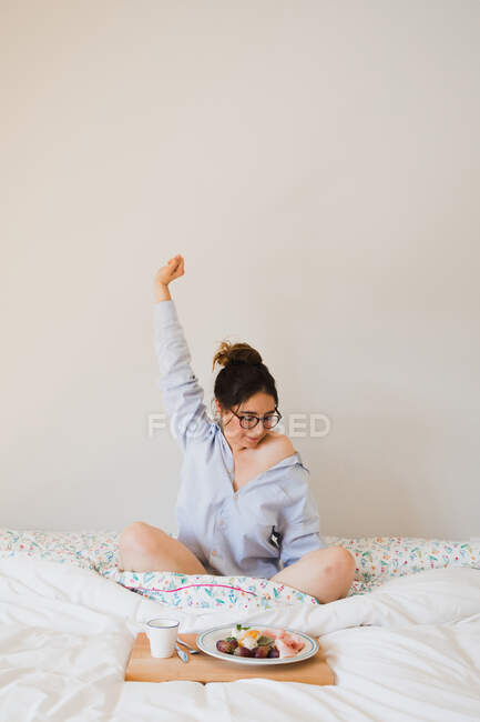 Porträt einer Frau, die sich ausstreckt, während sie auf dem Bett vor einem Tablett mit gesunder Nahrung sitzt — Stockfoto