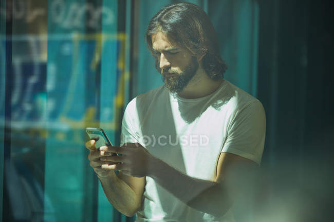 Junger bärtiger, gutaussehender Mann lehnt mit Handy in der Hand an der Wand und schreibt nachdenklich SMS — Stockfoto