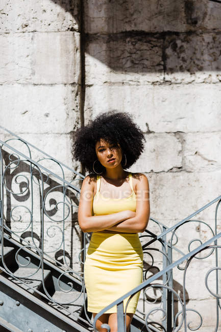 Афроамериканка в жёлтом платье, стоящая и опирающаяся на перила и смотрящая в камеру на городском фоне — стоковое фото