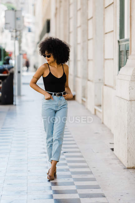 Jovem mulher étnica em jeans e top tanque andando ao ar livre — Fotografia de Stock