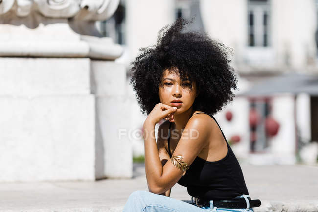 Donna etnica in jeans e canotta relax e prendere il sole su scale in pietra sullo sfondo urbano — Foto stock