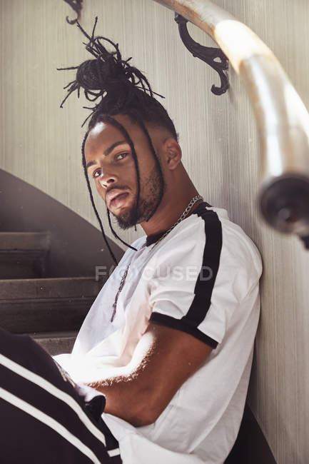 Афроамериканец с плетеными волосами сидит на лестнице и смотрит в камеру в помещении — стоковое фото
