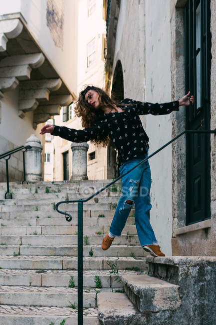 Joven mujer esbelta y casual estirándose y bailando en la escalera mientras baila con gracia en la calle de la ciudad vieja - foto de stock