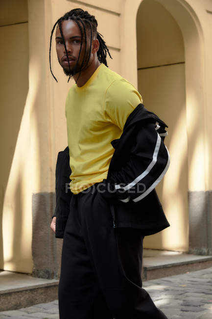 Joven hombre étnico con el pelo trenzado con traje deportivo negro mirando a la cámara en el fondo urbano - foto de stock