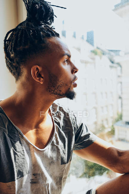 Задумчивый афроамериканец с косичками сидит дома на подоконнике — стоковое фото